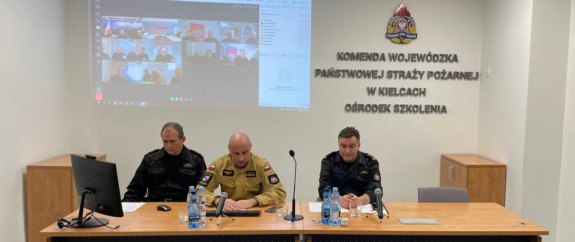 Zdjęcie przedstawia stół prezydialny, za którym siedzą: zastępca świętokrzyskiego komendanta PSP, naczelnik Ośrodka Szkolenia oraz jego zastępca. Strażacy prowadzą naradę pionu szkolenia. Za strażakami na ścianie jest wyświetlany obraz z projektora, na którym widać uczestników narady. Narada odbywa się w sali narad Komendy Wojewódzkiej PSP w Kielcach.