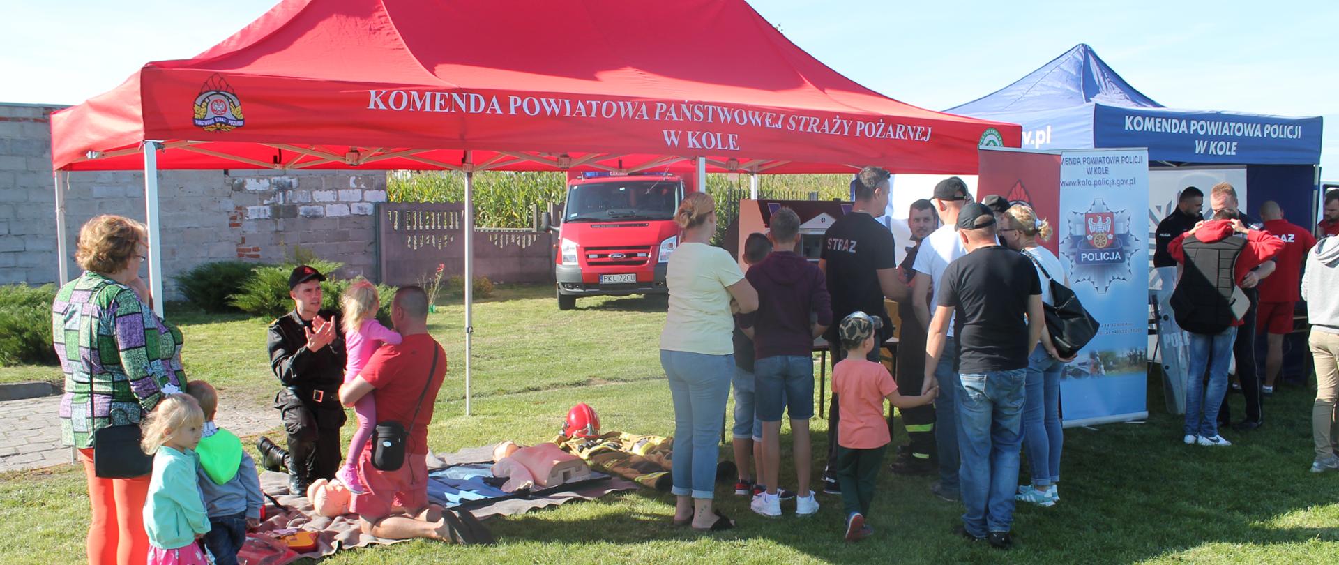 Zdjęcie przedstawia strażaka z Komendy Powiatowej Państwowej Straży Pożarnej w Kole udzielającego przybyłym na piknik osobom instruktażu pierwszej pomocy przedmedycznej oraz prezentującego mobilny symulator zagrożeń pożarowych domu jednorodzinnego