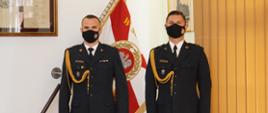 dwóch strażaków stoi na baczność w ciemnych mundurach, maja maseczki na twarzy, w tle sztandar