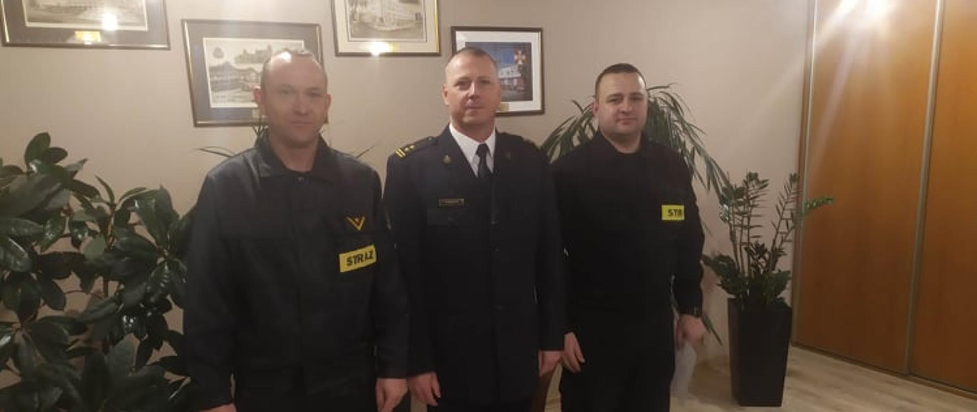 Na zdjęciu stoją dwaj funkcjonariusze w umundurowaniu koszarowym, w środku w umundurowaniu galowym komendant powiatowy PSP. za nimi na ścianie widać ryciny w ramach, na podłodze po lewej i prawej stronie stoją zielone kwiaty w dużych donicach. po prawej stronie widać szafę.