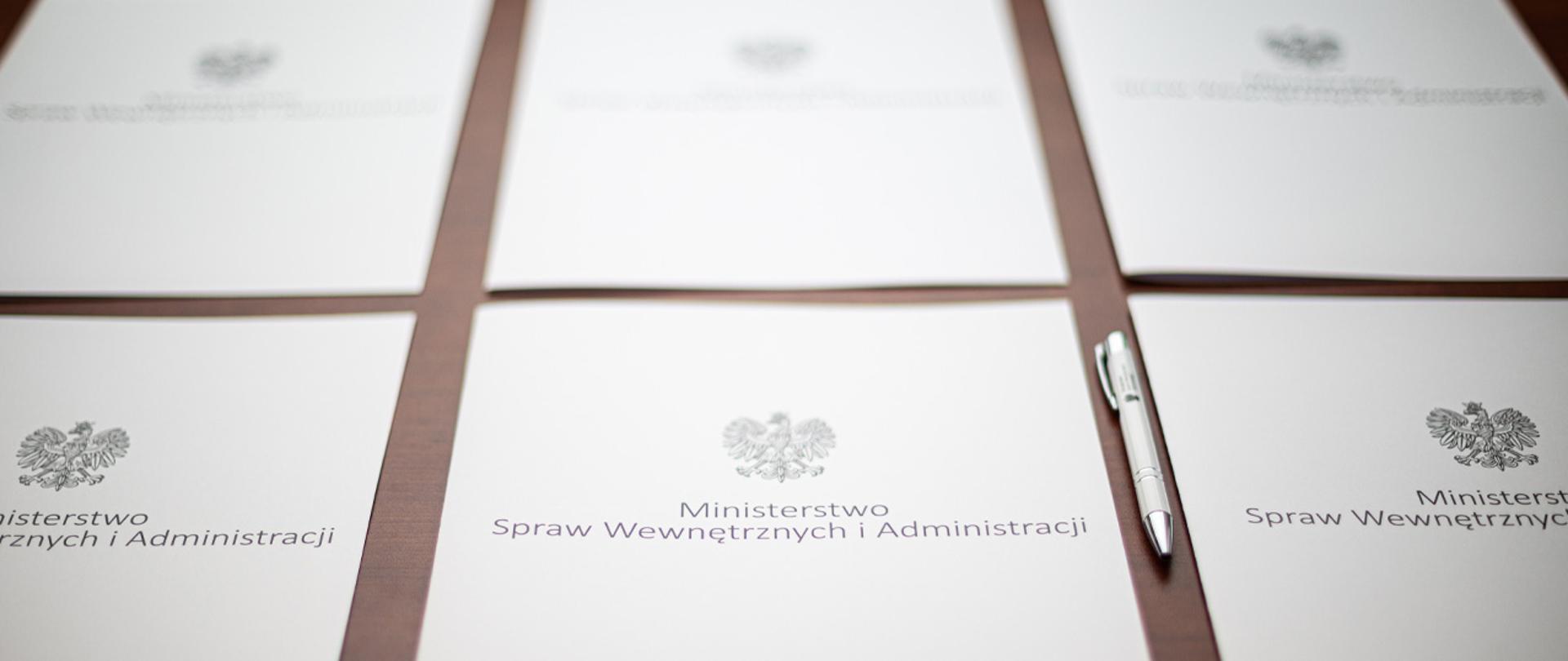 Na zdjęciu widać sześć leżących obok siebie teczek na dokumenty z napisem "Ministerstwo Spraw Wewnętrznych i Administracji" i orłem z godła narodowego. Obok jednej z nich leży długopis.