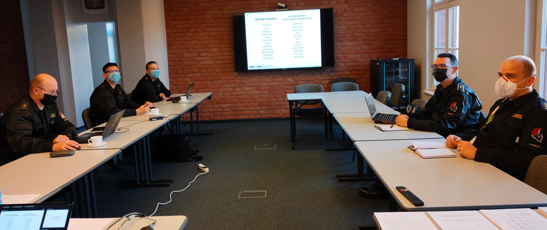 strażacy w maseczkach na twarzach siedzą przy stole, na ścianie wisi telewizor ze slajdem
