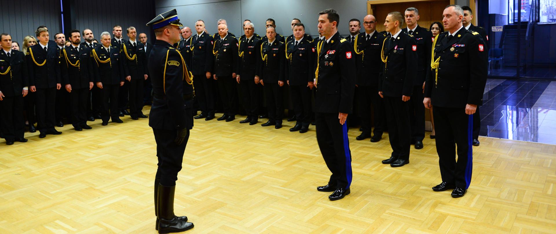 Widok z boku. Uroczysty apel. Dowódca uroczystości składa meldunek Komendantowi Głównemu Państwowej Straży Pożarnej, za Komendantem Głównym soją - od lewej przyjmujący obowiązki MKW, a następnie zdający obowiązki MKW
