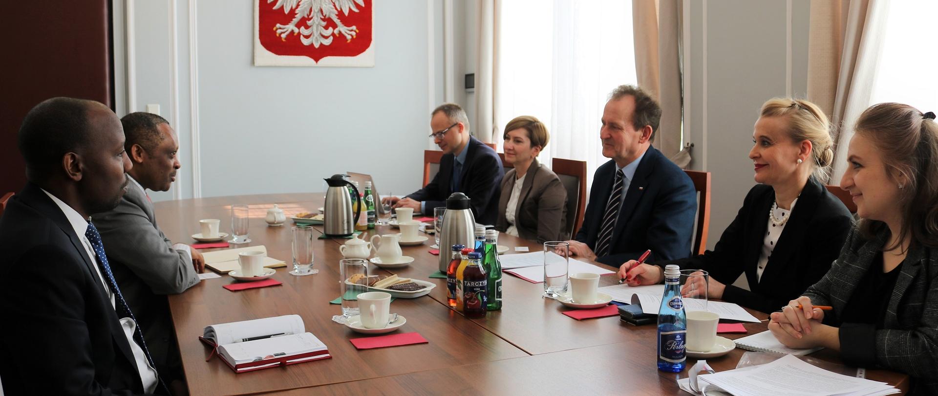 Zdjęcie zbiorowe - uczestnicy spotkania siedzą naprzeciwko siebie za stołem, za nimi ściana z godłem RP.