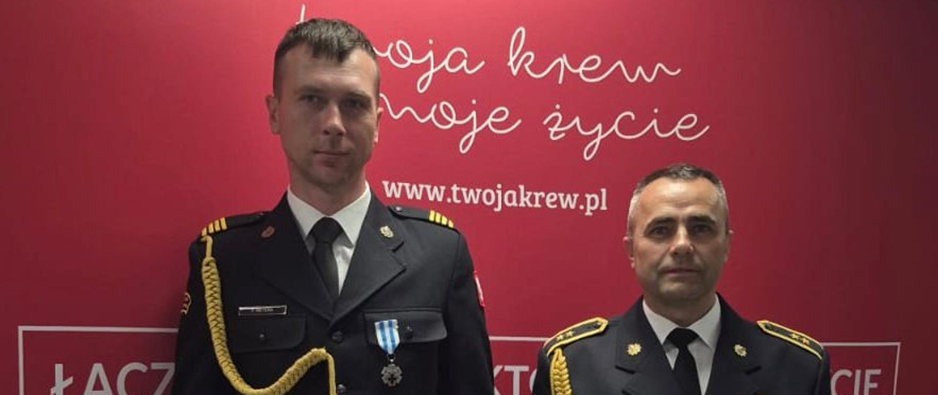 Na zdjęciu widocznych jest dwóch mężczyzn stojących obok siebie na tle czerwonej ścianki reklamowej. Obaj są ubrani w ciemne, wyjściowe mundury Państwowej Straży Pożarnej (PSP).