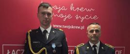 Na zdjęciu widocznych jest dwóch mężczyzn stojących obok siebie na tle czerwonej ścianki reklamowej. Obaj są ubrani w ciemne, wyjściowe mundury Państwowej Straży Pożarnej (PSP).
