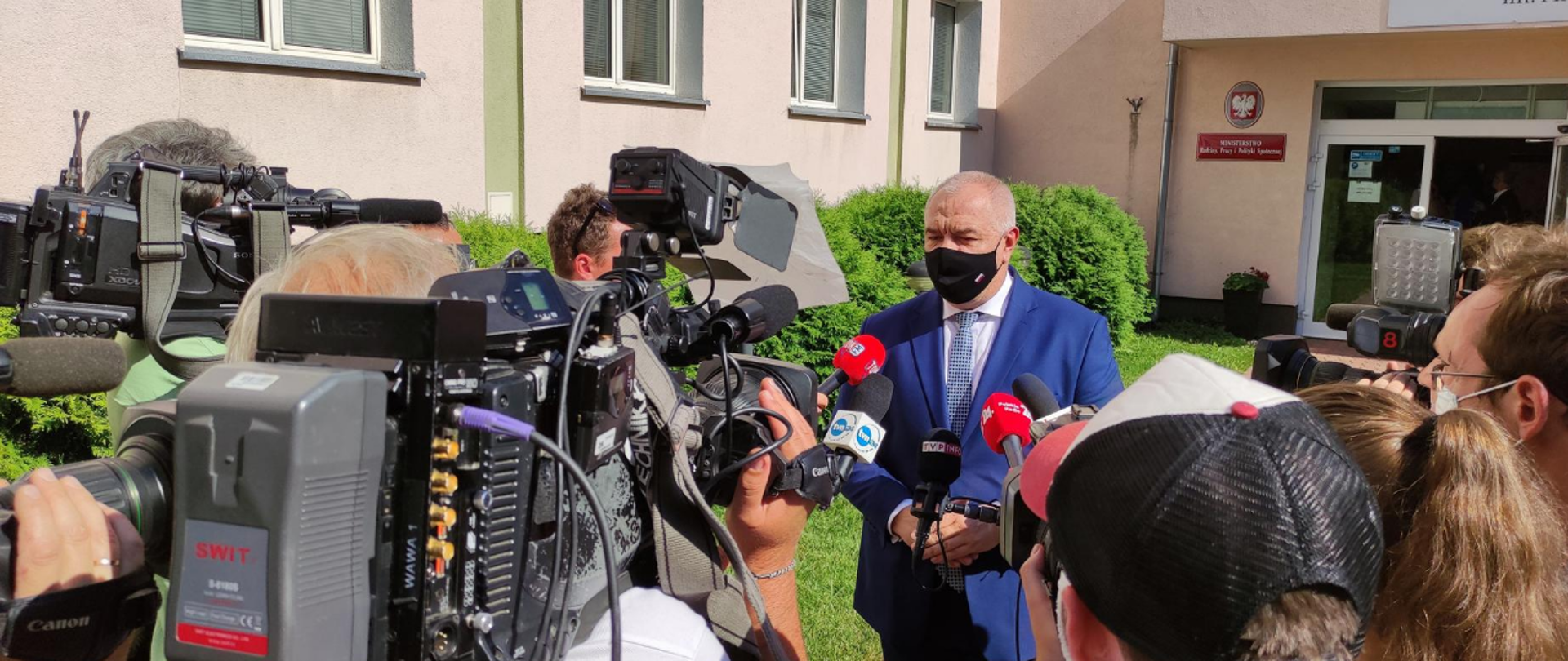 Wicepremier Jacek Sasin stoi przed budynkiem Centrum Partnerstwa Społecznego Dialog. Nos i usta zasłania mu czarna maseczka. Otaczają go dziennikarze i operatorzy telewizyjni. Na ujęciu widać mikrofony i kamery