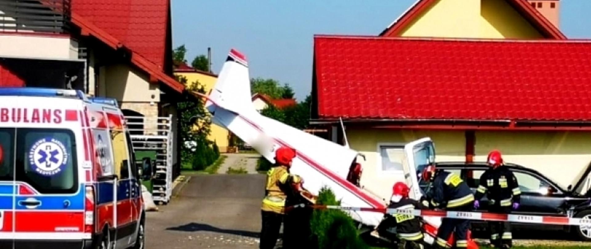Na zdjęciu znajduje się murowany jednorodzinny budynek mieszkalny z elewacją koloru jasnożółtego pokryty czerwoną blachodachówką. Przed budynkiem, na pierwszym planie widoczny jest mały biały samolot, zwijający na ogrodzeniu posesji w taki sposób, że jego ogon znajduje się w wysoko w powietrzu, po jednej zewnętrznej stronie ogrodzenia nad asfaltową drogą dojazdową do tej i innych posesji, a dziób oparty jest o zielony trawnik, po drugiej stronie ogrodzenia. Pomiędzy samolotem a budynkiem stoi, częściowo zasłonięty wrakiem samolotu, granatowy samochód osobowy z otwartą maską silnika, a wokół nich pracują trzej strażacy ubrani w ubrania specjalne., którzy wykonują czynności ratownicze przy otwartej kabinie pilota maszyny. Jeden (czwarty) strażak w ubraniu specjalnym stoi po zewnętrznej stronie ogrodzenia na skraju asfaltowej drogi za pomocą czerwono – białej taśmy wygradza strefę prowadzonych działań. Przed budynkiem, po lewej stronie fotografii na ulicy stoi zaparkowana karetka pogotowia ratunkowego. Zdjęcie wykonano w bardzo dobrych warunkach pogodowych – u góry fitografii widoczne jest błękitne, bezchmurne niebo.