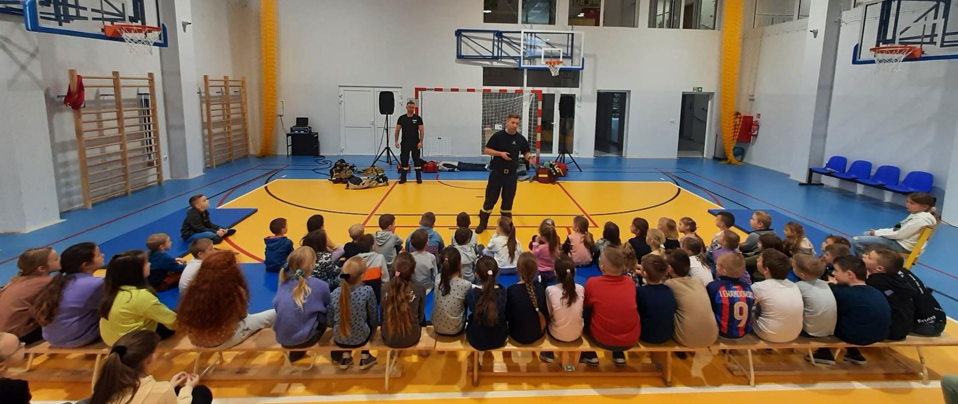 Zdjęcie wykonane na sali gimnastycznej Publicznej Szkoły Podstawowej w Cerekwi. Na zdjęciu widać dwóch strażaków. Jeden z nich stoi przed siedzącymi na ziemi dziećmi – uczniami szkoły podstawowej. Za nim w pewnej odległości stoi drugi. Obok niego na ziemi leży umundurowanie specjalne i ochronne strażaka używane podczas działań ratowniczo – gaśniczych, manekin do prezentacji zasad udzielania pierwszej pomocy oraz torba medyczna w której znajduje się niezbędny asortyment przeznaczony do udzielania pierwszej pomocy. 