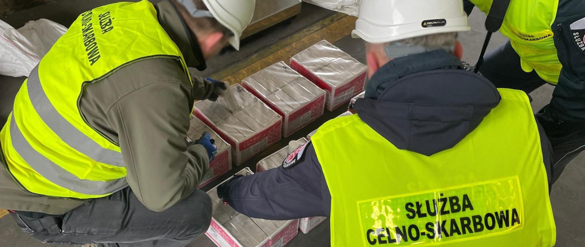 Funkcjonariusze Służby Celno‑Skarbowej w kamizelkach odblaskowych i kaskach kontrolują kartony z towarem w magazynie.