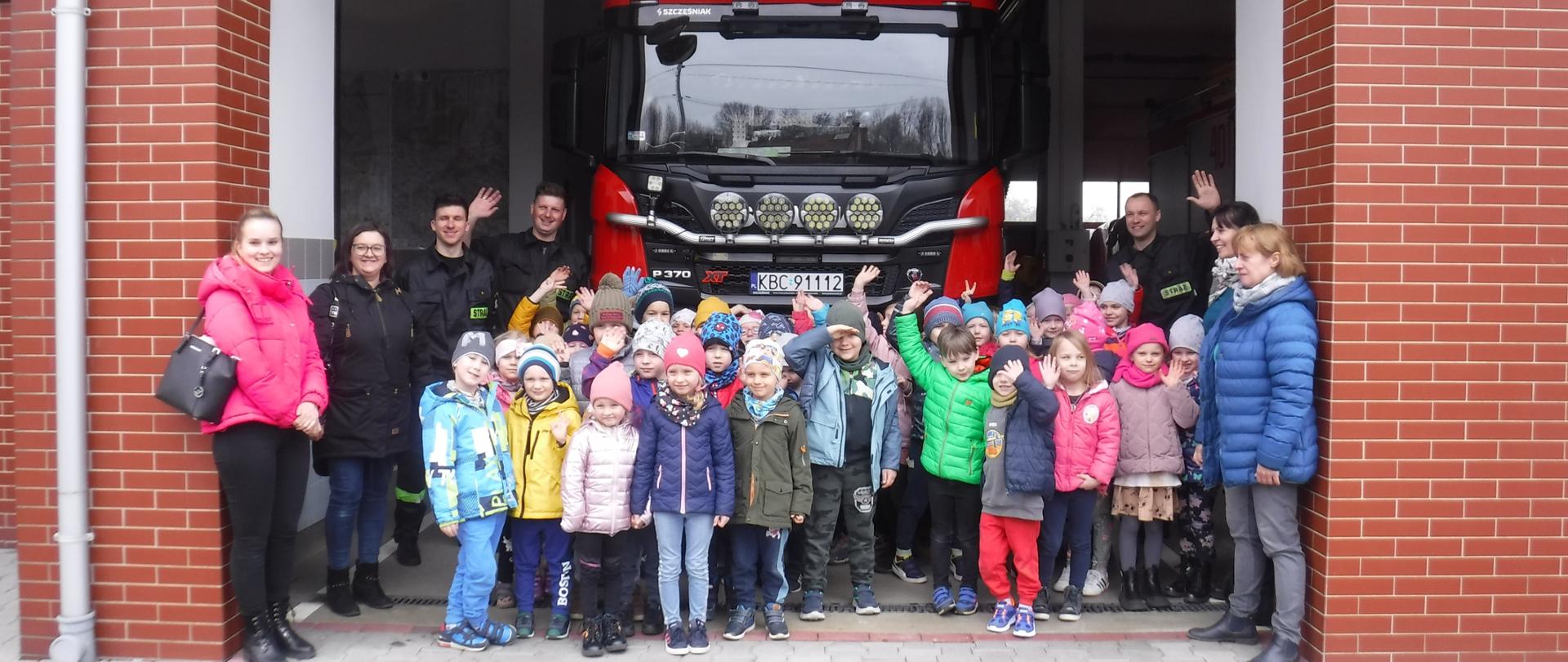 Kilkadziesiąt dzieci ubranych w kurtki i czapki, stoją na kostce betonowej przed bramami garażowymi oznaczonymi czerwonymi numerami. W tle budynek Komendy Powiatowej Państwowej Straży Pożarnej w Bochni. Budynek w kolorze białym, okna i drzwi koloru białego