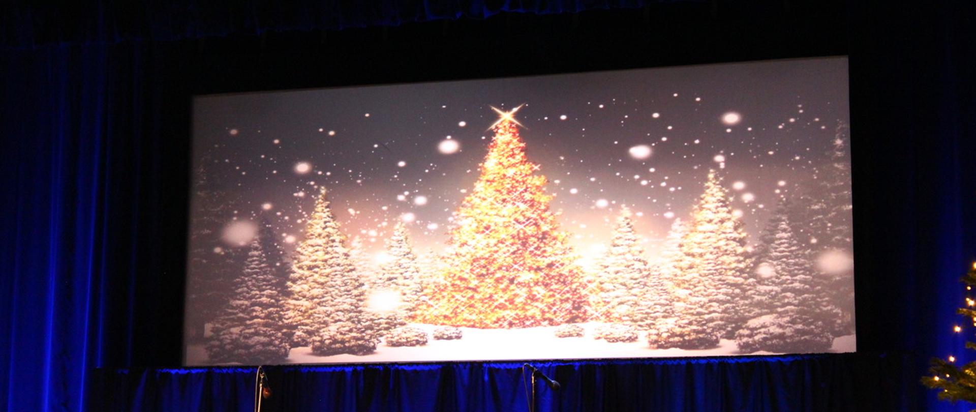 na scenie wyświetlany jest plakat z projektora, ukazujący świecące choinki w zimowej aurze, w tle ciemno-niebieskie kotary oraz choinka z włączonymi lampkami