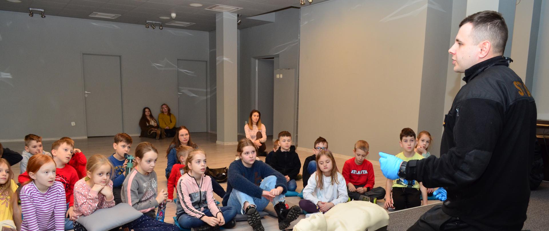 Dzieci obserwują i słuchają strażaka, który prezentuje zasady udzielania pomocy z wykorzystaniem fantoma.