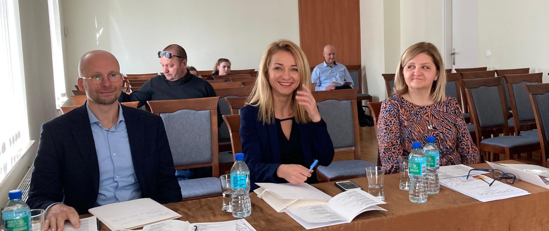 Sala koncertowa Państwowej Szkoły Muzycznej I st. im. K. Namysłowskiego w Szczebrzeszynie. Za stołem przykrytym brązowym materiałem siedzą jurorzy IX Konkursu Gitarowego - od prawej - sekretarz Beata Płocharz, po środku Ewa Jabłczyńska, po prawej stronie Dariusz Kupiński. Na stole leżą kartki i programy. Po prawej stronie na widowni siedzi pani, z tyłu na widowni siedzi kilka osób. 