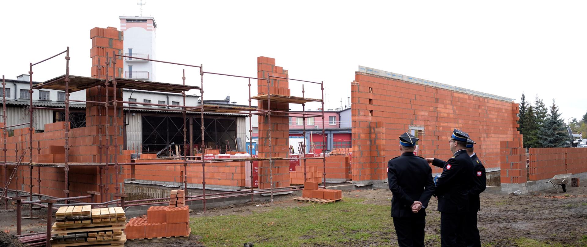 Na zdjęciu widać plac budowy. Mury wymurowane z czerwonej cegły. W centralnej części fotografii trzech funkcjonariuszy PSP w umundurowaniu. Zdjęcie wykonane w ciągu dnia. Na zdjęciu widać również rośliny zielone oraz sprzęt budowlany.