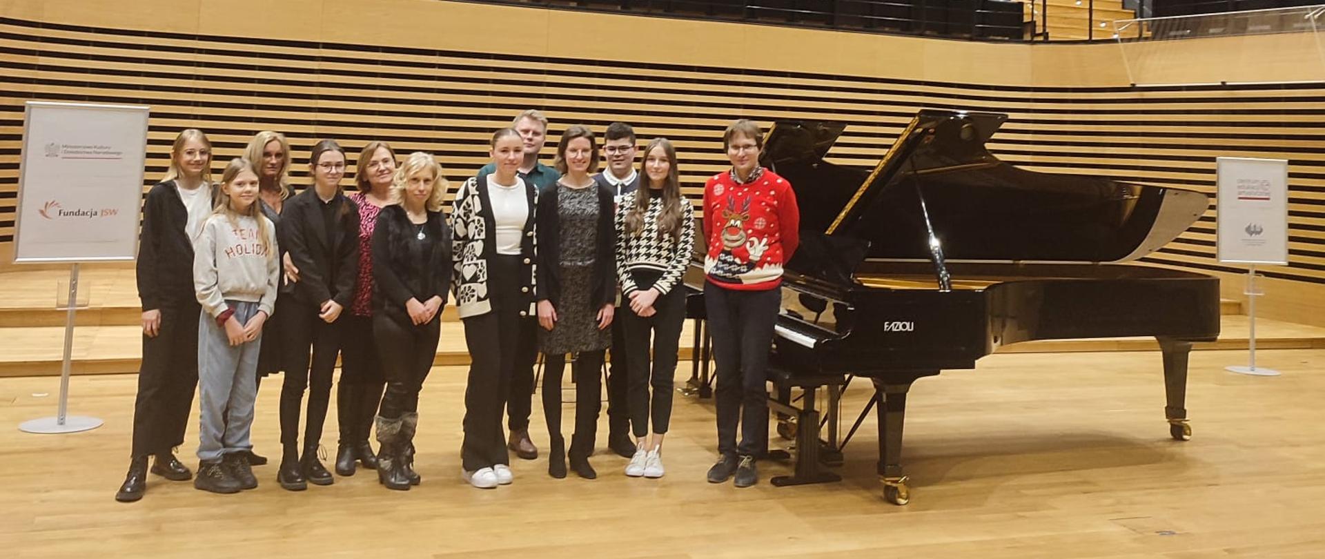 Grupa uczestników warsztatów (sześciu uczniów i pięciu nauczycieli) oraz Krzysztof Książek obok fortepianu Fazioli na scenie sali koncertowej PSM