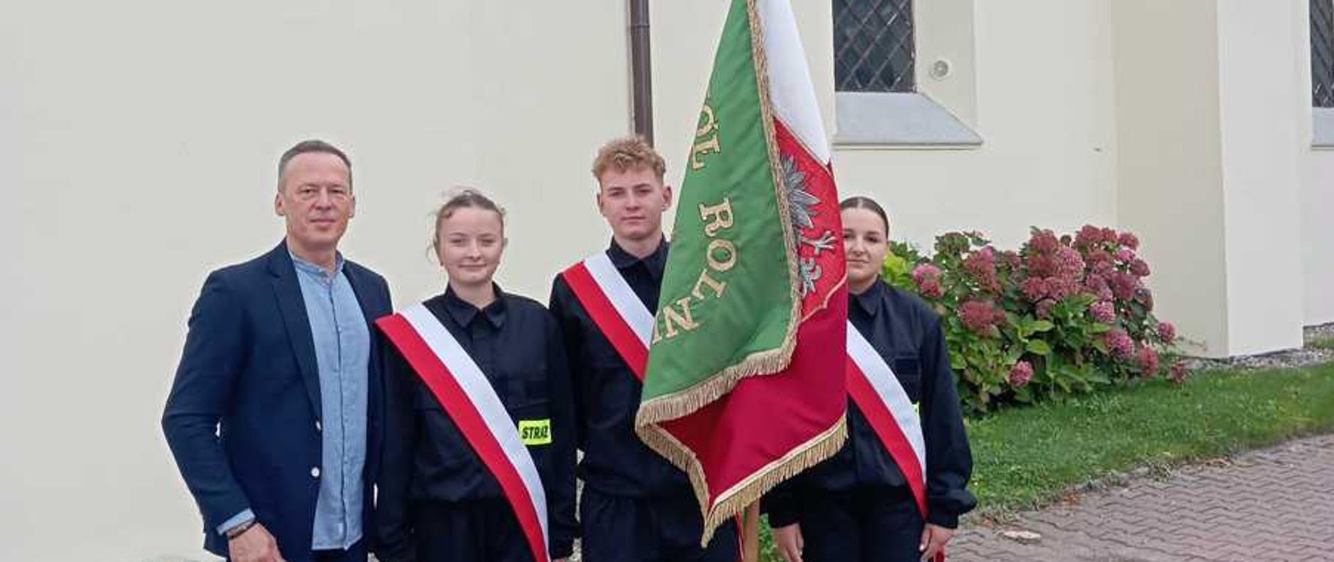 Zdjęcie przedstawia cztery osoby stojące na brukowanym chodniku przed jasnym, klasycznym budynkiem sakralnym lub zabytkowym (prawdopodobnie kościołem).Od lewej strony stoją:Mężczyzna w wieku średnim – ubrany w elegancką, ciemnoniebieską marynarkę, ciemną koszulę, klasyczne jeansy oraz czarne buty sportowe. Uśmiecha się w stronę obiektywu.Młoda kobieta w mundurze – ma na sobie czarny strój przypominający mundur strażacki (z widocznym fragmentem napisu „STRAŻ” na żółtym tle) oraz wysokie buty robocze z żółtymi elementami. Przez ramię ma przewieszoną biało-czerwoną szarfę.Młody mężczyzna (poczet sztandarowy) – ubrany w taki sam czarny mundur i wysokie buty. Przez ramię ma założoną biało-czerwoną szarfę, a w dłoniach trzyma duży sztandar. Widoczny fragment napisu na zielonym tle sztandaru to „Ł ROLN...”, a obok znajduje się godło Polski (orzeł w koronie na czerwonym tle). Sztandar wykończony jest złotymi frędzlami.Druga młoda kobieta – również ubrana w identyczny czarny mundur strażacki, wysokie buty oraz biało-czerwoną szarfę.Tło zdjęcia stanowi jasna, kremowa ściana budynku z podłużnymi, łukowatymi oknami. Po prawej stronie, wzdłuż ściany, rosną zielone krzewy z różowymi kwiatami (hortensje), a za nimi rozciąga się trawnik. Całość grupy oświetlona jest naturalnym, dziennym światłem.