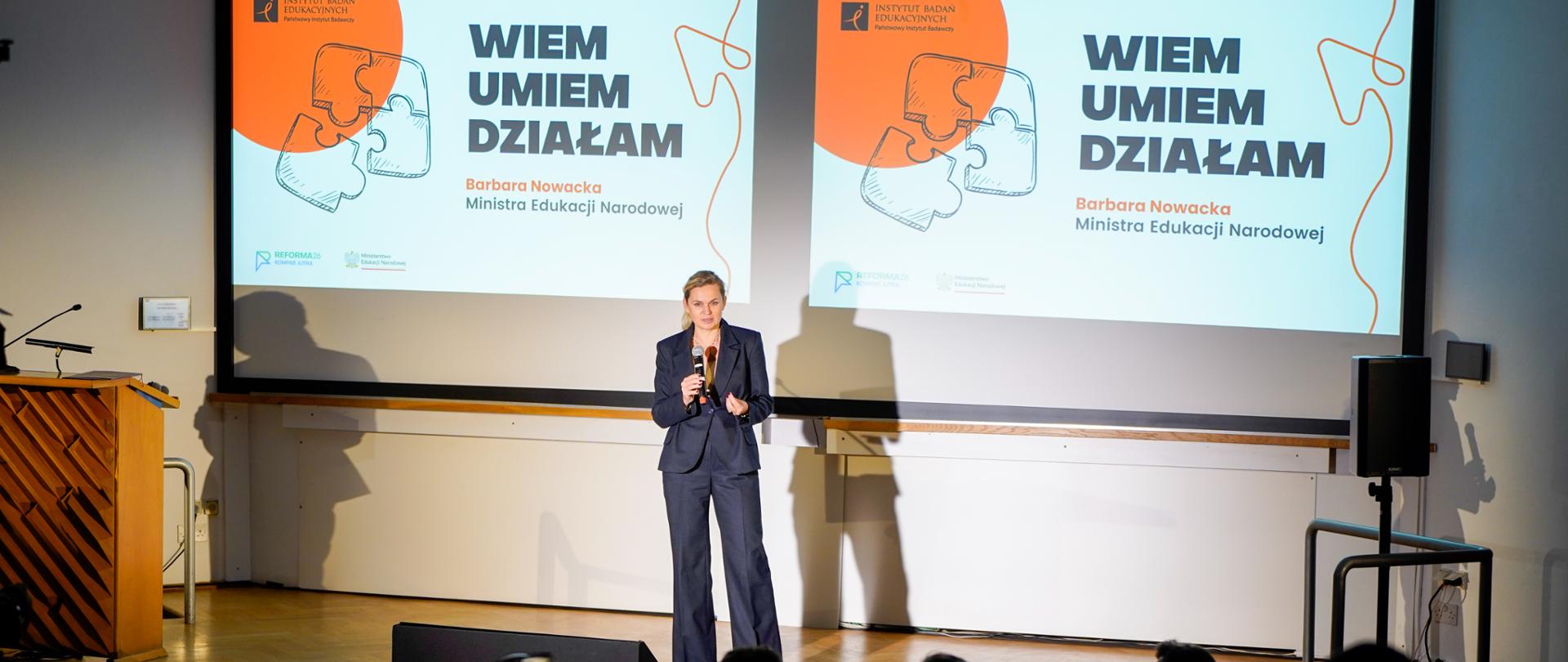 Szeroki plan z tyłu widowni. Barbara Nowacka, Minister Edukacji Narodowej, stoi na scenie i przemawia do widowni, której sylwetki są widoczne na pierwszym planie w półmroku. W tle za prelegentką dwa duże ekrany z tytułem: „WIEM UMIEM DZIAŁAM” i jej nazwiskiem oraz stanowiskiem.