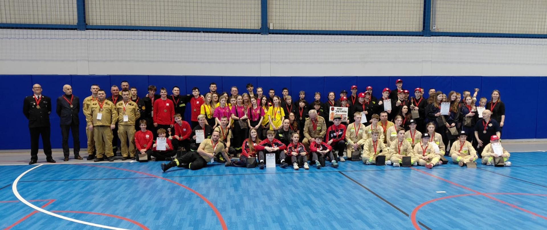 Szerokie zdjęcie pamiątkowe wszystkich uczestników, organizatorów i sędziów zawodów, stojących i klęczących na niebieskiej podłodze hali sportowej. W centrum znajduje się duża grupa młodzieży w różnych mundurach, wielu z nich ma medale na szyjach i dyplomy w rękach. Po bokach stoją sędziowie i oficjele w mundurach galowych. W tle widoczna jest duża niebieska ściana i trybuny.