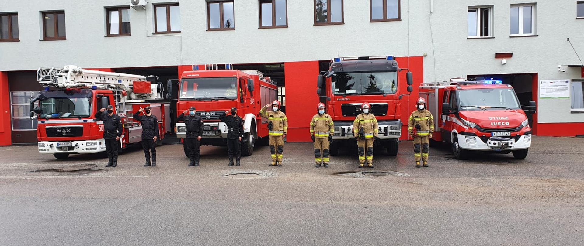 Strażacy stoją na placu Komendy przed samochodami strażackimi z włączonymi sygnałami świetlnymi i dźwiękowymi w celu uczczenia poległego na służbie Policjanta. 