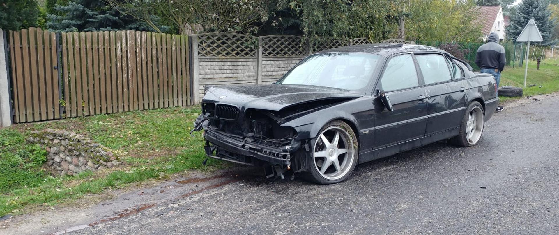 Samochód osobowy marki BMW stoi częściowo na poboczu i na drodze. Ma wgnieciony przód. Za nim leży koło, które odpadło podczas zderzenia. 