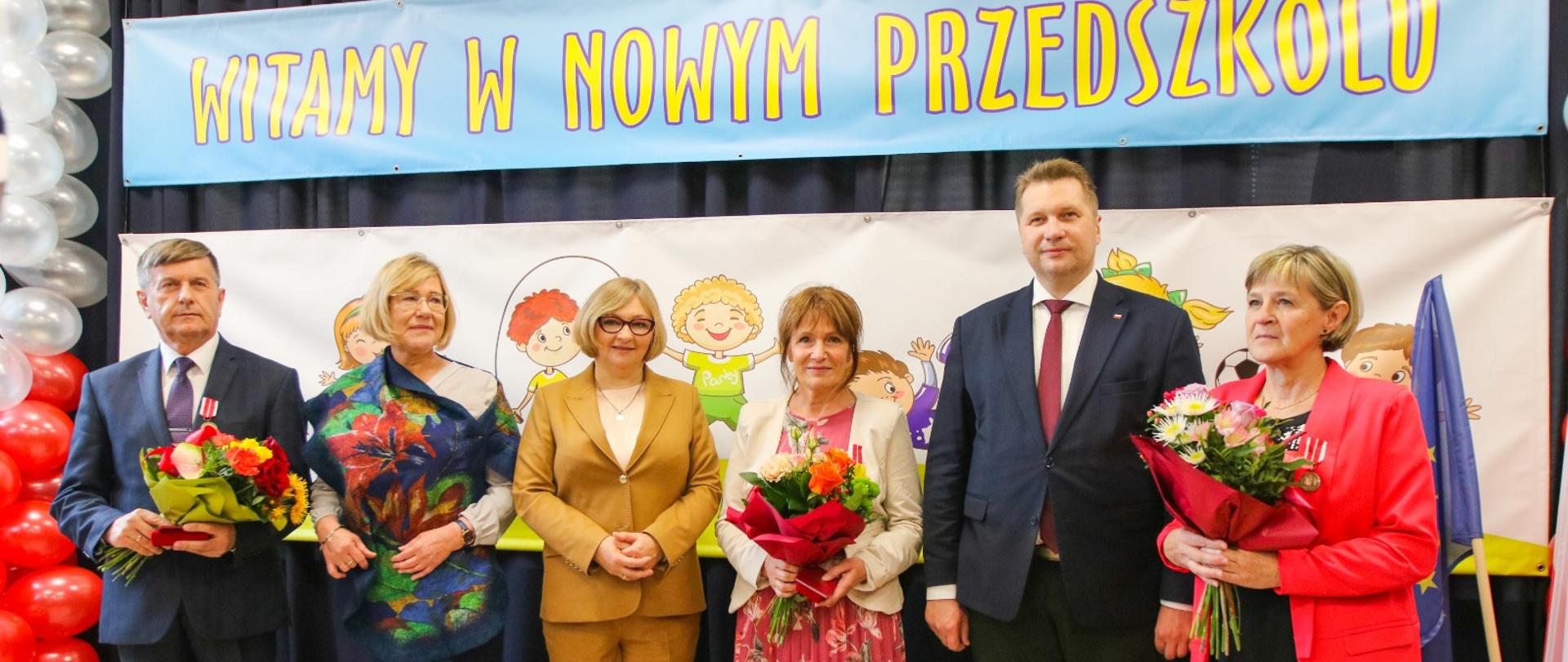 Minister Czarnek w otoczeniu grupy kolorowo ubranych osób stoi pod ścianą z napisem Witamy w nowym przedszkolu.