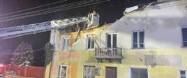 Widoku zniszczonego dachu i stropu nad kamienica. Strażacy z kosza drabiny mechanicznej przeszukują budynek przy użyciu kamery termowizyjnej. Pora nocna . 