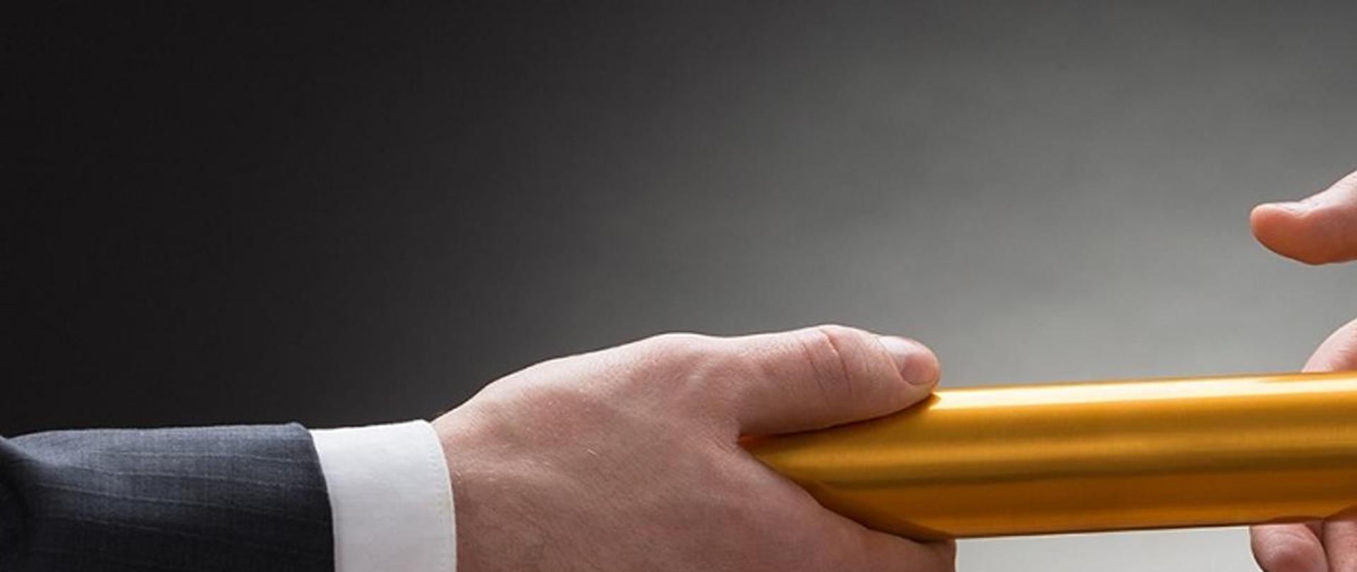 Grafika przedstawiająca dwie męskie dłonie, które przekazują sobie dokument