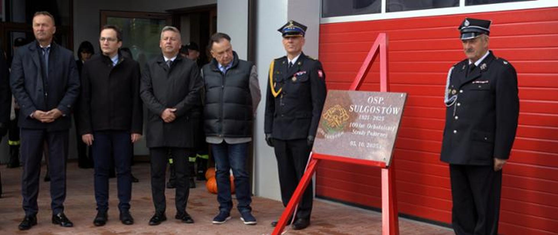 Zdjęcie przedstawia grupę mężczyzn stojących przed czerwonymi, rolowanymi drzwiami garażowymi straży pożarnej, obok pamiątkowej tablicy.
Po lewej stronie znajduje się czterech mężczyzn ubranych w cywilne płaszcze i kurtki. Dwóch z nich (pierwszy i trzeci od lewej) ma ciemne garnitury i płaszcze, drugi (od lewej) jest w czarnym płaszczu, a czwarty (od lewej) w granatowej, pikowanej kamizelce na swetrze i dżinsach. Wszyscy stoją na wprost, patrząc w stronę aparatu, z poważnymi minami.
Po prawej stronie stoją dwaj mężczyźni w galowych mundurach strażackich, z czapkami na głowach, również patrzący w stronę aparatu.
Między obiema grupami znajduje się ciemna, prostokątna tablica umieszczona na metalowym stojaku. Na tablicy, ozdobionej herbem, widnieje napis w języku polskim:
OSP SULGOSTÓW
1925 - 2025
100-lecie Ochotniczej Straży Pożarnej
05.10.2025 r.
W tle widać fragmenty budynku strażnicy oraz kolejnych ludzi w mundurach w głębi po lewej.