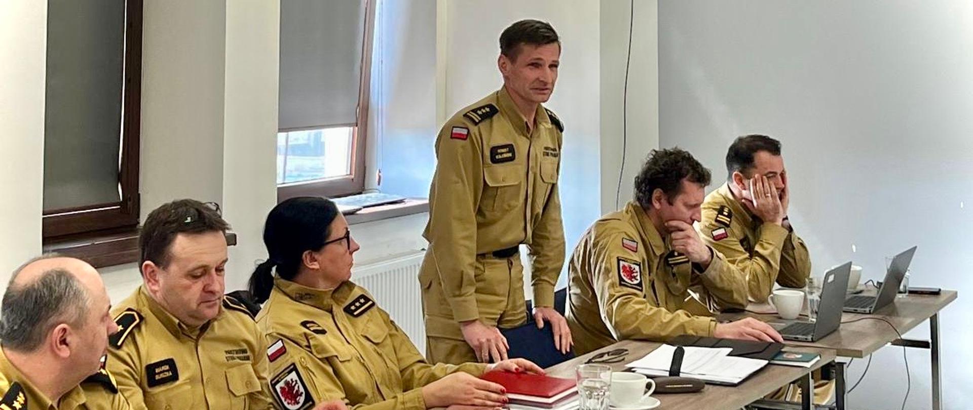 Za stołem sześcioro strażaków w mundurach służbowych, po środku jeden z nich stoi i przemawia. Na stołach są dokumenty, laptopy, naczynia... W tle okna sali. 