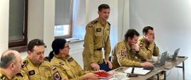 Za stołem sześcioro strażaków w mundurach służbowych, po środku jeden z nich stoi i przemawia. Na stołach są dokumenty, laptopy, naczynia... W tle okna sali. 