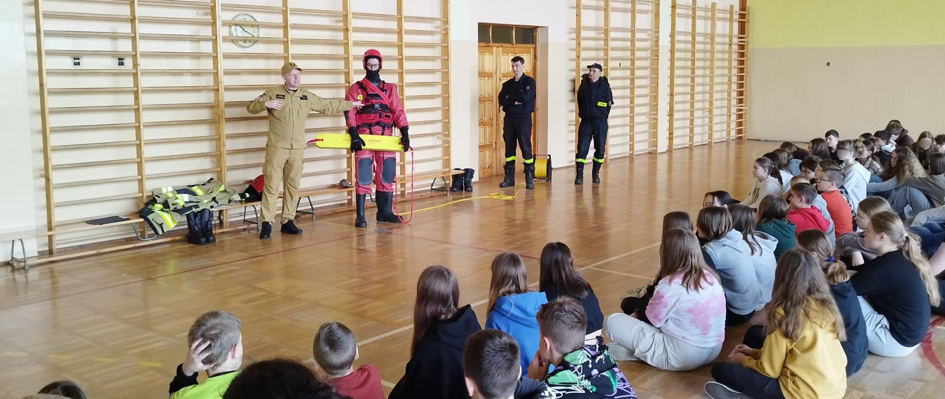 Zdjęcie przedstawia dzieci siedzące na podłodze sali gimnastycznej dalej strażacy prezentują sprzęt wykorzystywany podczas działań ratowniczych na akwenach wodnych.