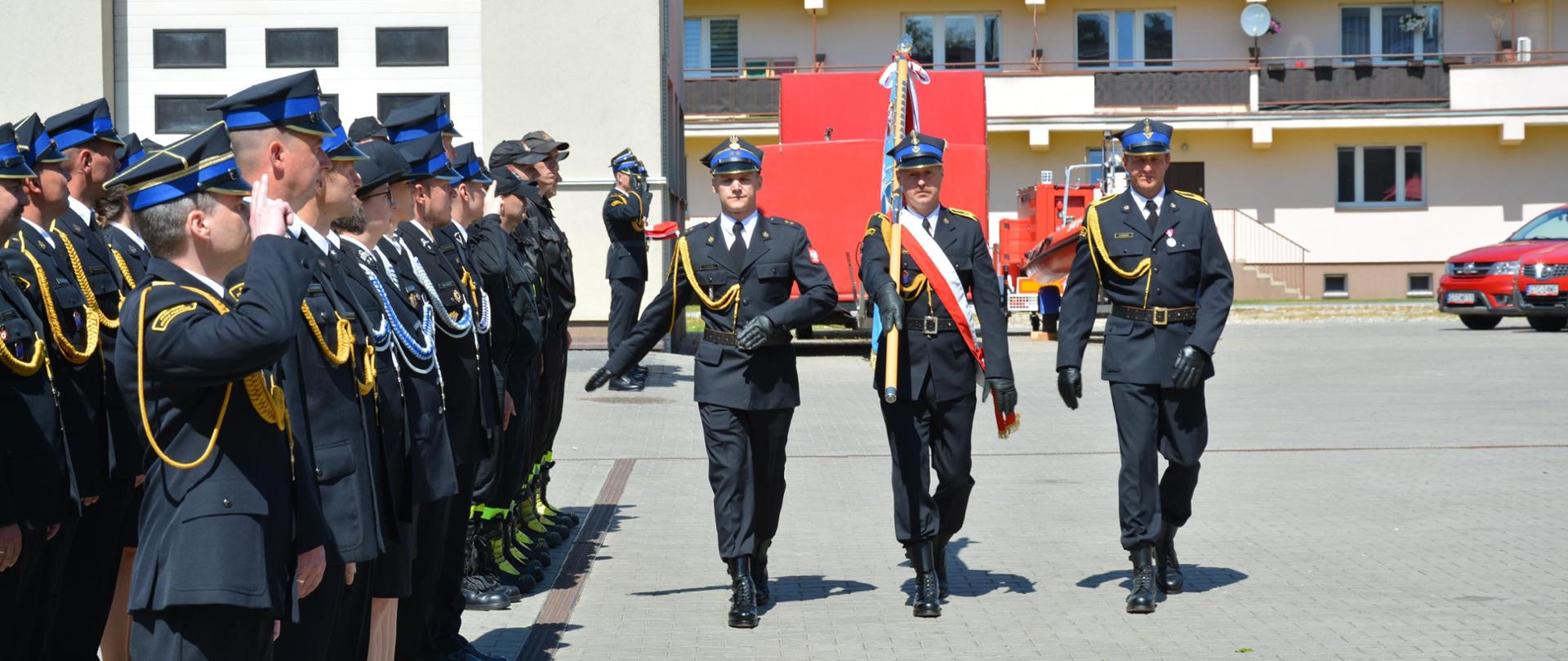Poczet sztandarowy wprowadza sztandar KP PSP Tczew idą wzdłuż pododdziału złożonego ze strażaków w mundurach wyjściowych.
