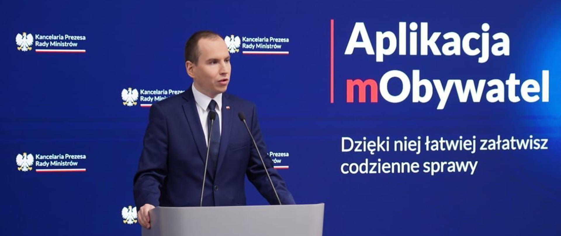 Na zdjęciu minister Adam Andruszkiewicz podczas konferencji prasowej w Kancelarii Premiera. Na ekranie w tle wyświetlany jest tekst "Aplikacja mObywatel. Dzięki niej łatwiej załatwisz codzienne sprawy" 