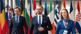 Minister Marcin Kulasek idzie w towarzystwie kobiety i mężczyzny. Wszyscy są elegancko ubrani. W tle widać flagi wielu państw.
