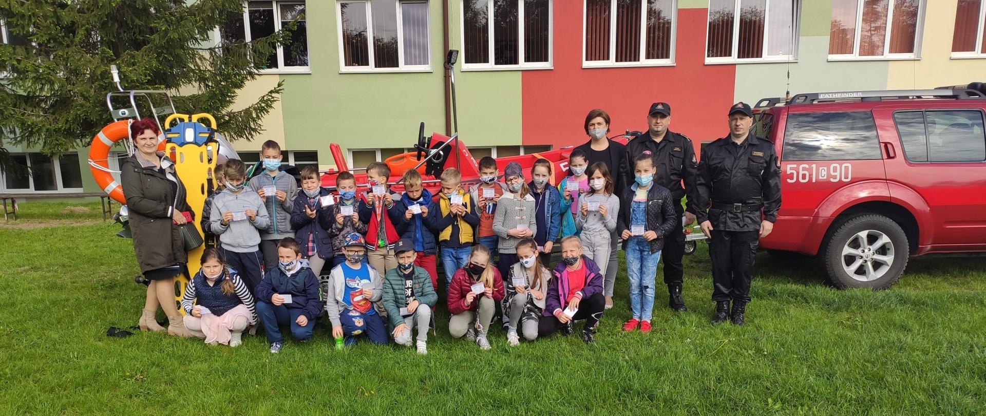 To zdjęcie grupowe dzieci, które uczestniczyły w szkoleniu. Obok dzieci dwóch strażaków stoi w umundurowaniu sztabowym, za plecami ustawiono pojazd strażacki z łodzią na przyczepie. Wszyscy są na tle budynku szkoły.