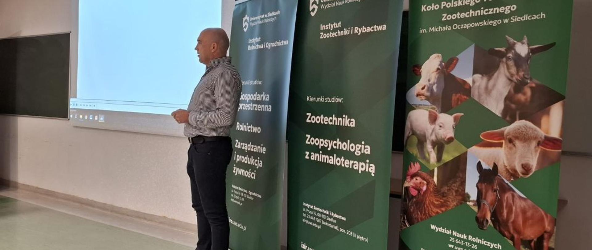 Prowadzący wykład mężczyzna stoi obok dużego ekranu, za nim roll-upy o tematyce rolniczej i zootechnicznej 