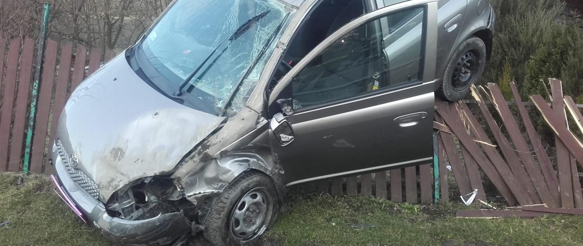 Samochód osobowy z uszkodzonym przodem zawieszony na drewnianym płocie. 