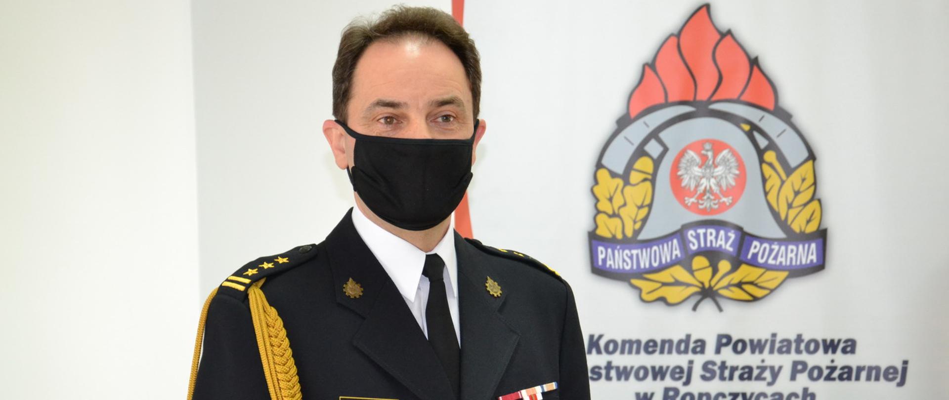 st. bryg. Janusz Piczak Z-ca Komendanta Powiatowego w mundurze galowym na tle logo PSP