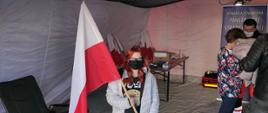 Widok z przodu. W namiocie – mobilnym punkcie szczepień kobieta w maseczce ochronnej trzyma w rękach biało-czerwoną flagę otrzymaną za przystąpienie do akcji „Zaszczep się dla Niepodległej”. 