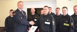 Zdjęcie przedstawia moment gratulacji dla wyróżnionego strażaka. Komendant Powiatowy wręcza dokument. W tle zmiana służbowa