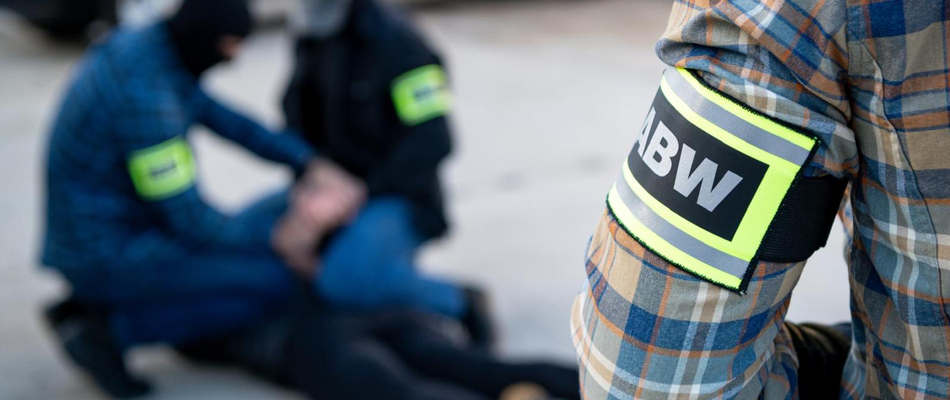 Po prawej stronie zdjęcia widać ramię funkcjonariusza ABW. Funkcjonariusz jest ubrany w koszulę w kratkę (kolory: szary, niebieski, biały, brązowy i pomarańczowy). Na przedramieniu funkcjonariusza widać odblaskowe oznaczenie z napisem ABW. Po lewej stronie zdjęcia widać dwóch funkcjonariuszy ABW w kominiarkach i z odblaskowymi oznaczeniami, zatrzymujących osobę. Osoba zatrzymana leży na ziemi i są jej zakładane kajdanki. U góry zdjęcia widać również fragment ciemnego samochodu.