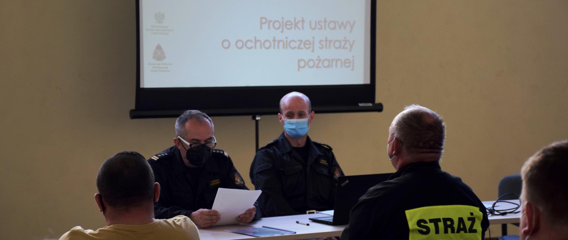 przodem do widza, za stołem prezydialnym siedzi dwóch strażaków w ubraniach dowódczo - sztabowych. Z tyłu ekran do wyświetlania prezentacji, na pierwszym planie strażacy siedzący tyłem do widza.