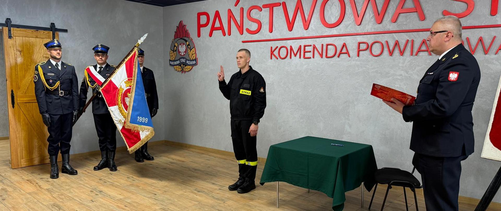 Strażaka który ślubuje. Stoi on w postawie zasadniczej z ręką w górze. Z prawej strony stoi komendant, który czyta rotę ślubowania. Z lewej strony stoi poczet sztandarowy. 