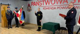Strażaka który ślubuje. Stoi on w postawie zasadniczej z ręką w górze. Z prawej strony stoi komendant, który czyta rotę ślubowania. Z lewej strony stoi poczet sztandarowy. 