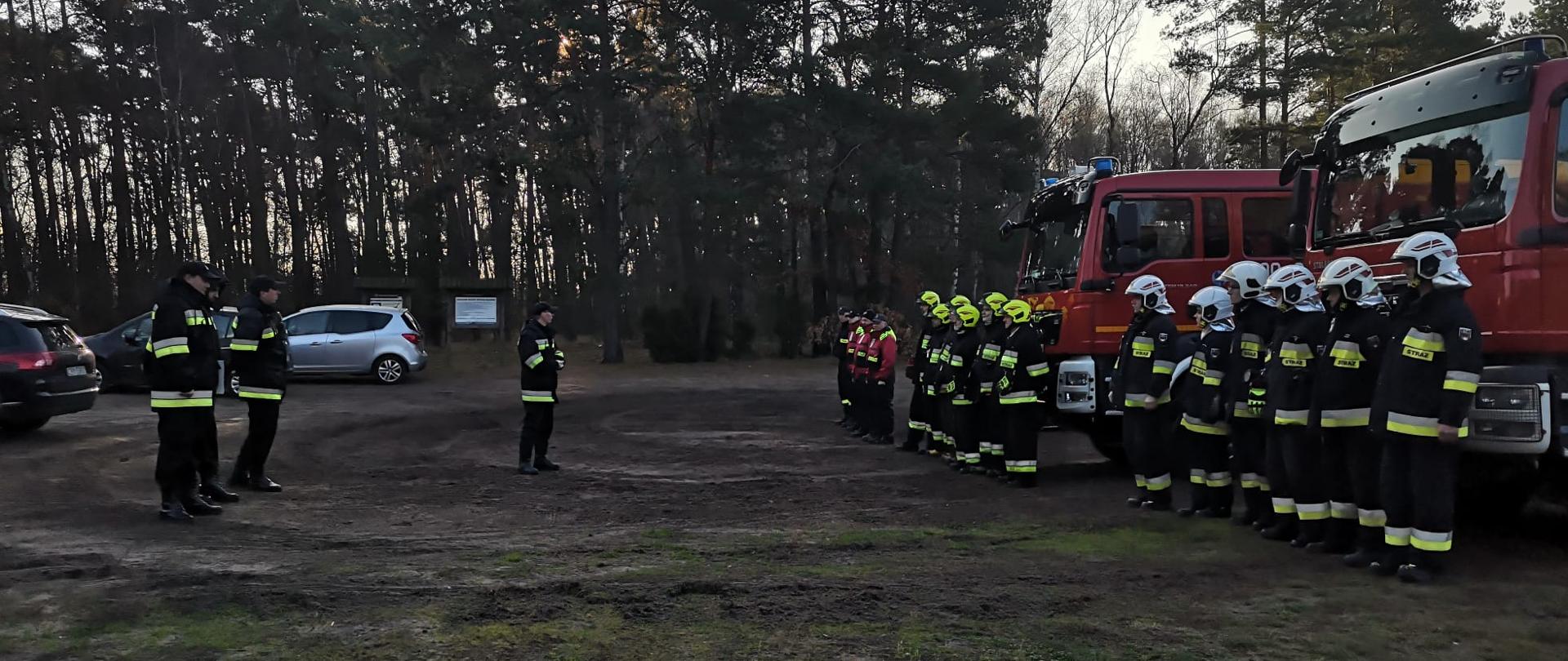Po prawej stronie zbiórka strażaków w szeregu przy samochodach. Po lewej stoją 4 osoby w ubraniach kadry dowódczo-sztabowej organizujące ćwiczenia. Jedna z tych osób wysunięta w stronę strażaków stojących w szeregu. 