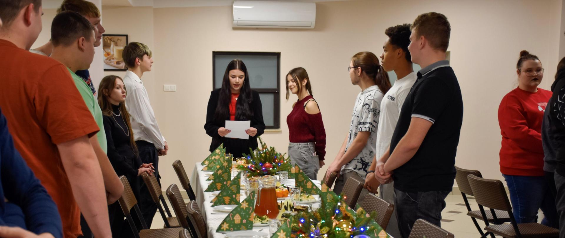Młodzież stoi przy zastawionym świątecznie stole, u szczytu uczennica czyta fragment biblii