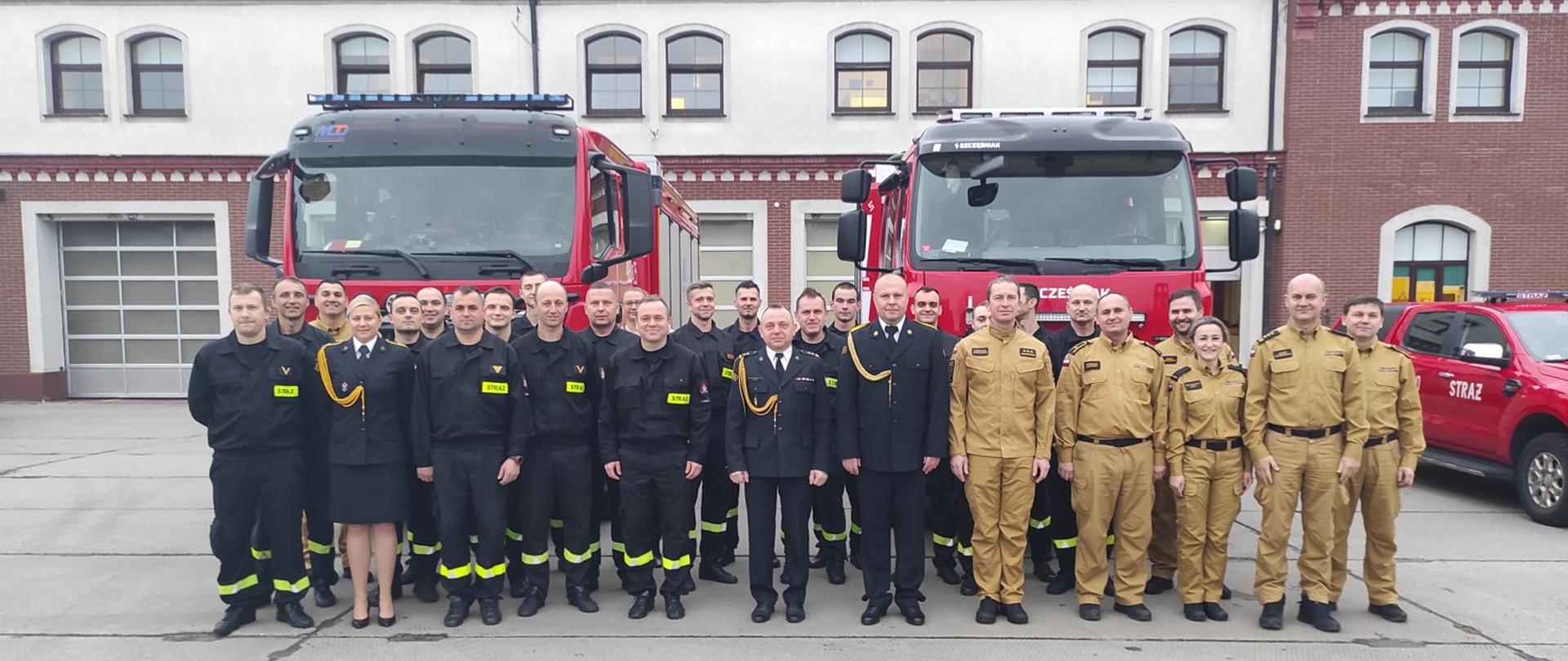Zdjęcie przedstawia strażaków stojących przed samochodami pożarniczymi, w tle budynek strażnicy