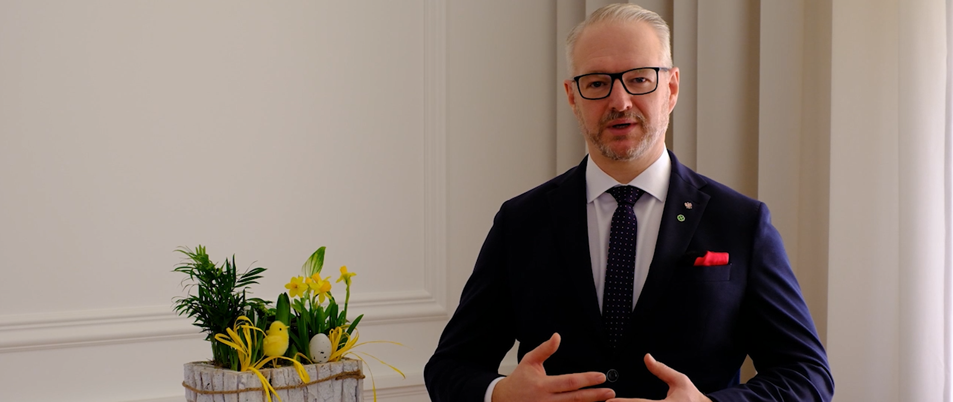 mężczyzna w garniturze gestykuluje, mówi do kamery, obok stoi stroik wielkanocny 