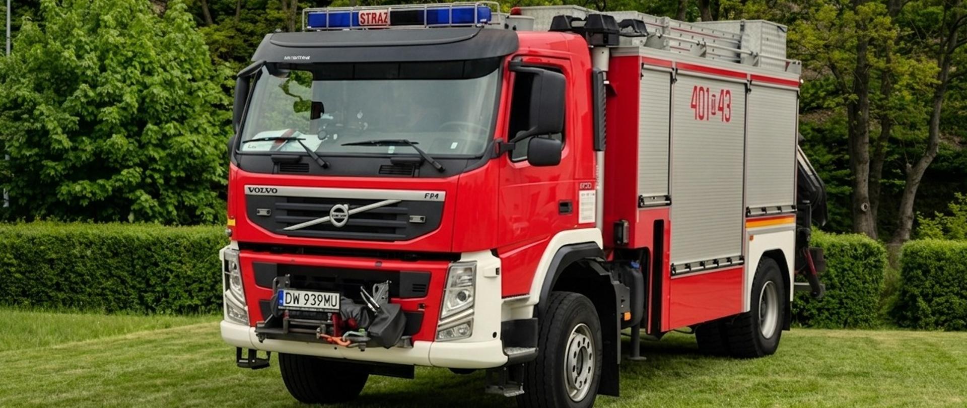 Ciężki samochód ratownictwa technicznego Volvo FM. Czerwone nadwozie z numerem na szarej skrytce „401[D]43” wyposażone jest w hydrauliczny dźwig żurawiowy za zabudową. Pojazd stoi na zielonej trawie w tle widać las liściasty.